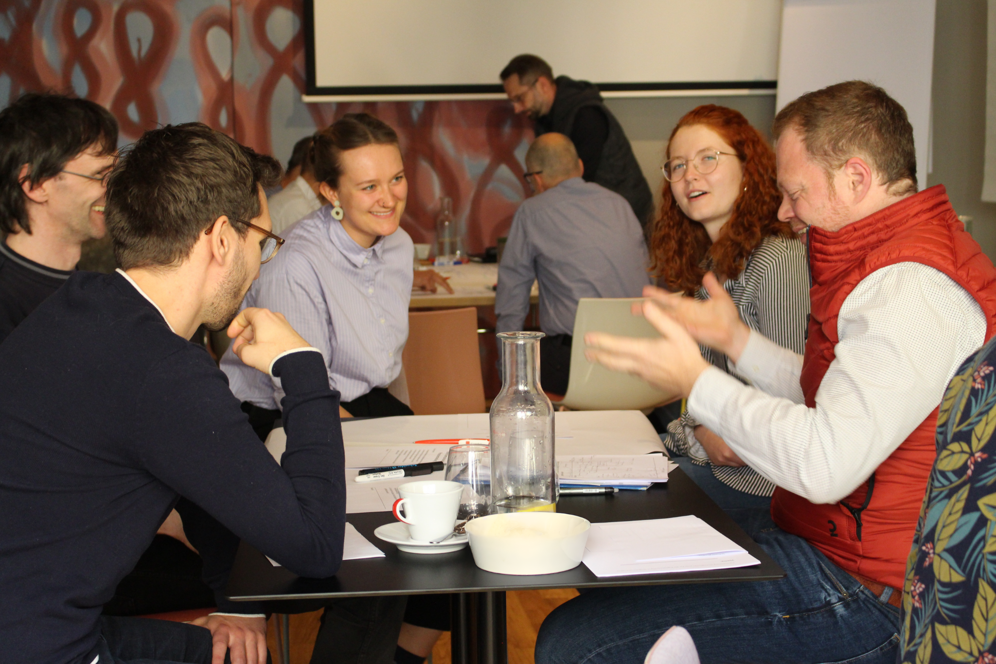 Ein Team von Lehrkräften und Mitarbeitern sitzt an einem Tisch in einer lebhaften Diskussionsrunde. Alle Teilnehmer lachen herzlich, und die Atmosphäre ist positiv und engagiert. Sie befinden sich in einem intensiven Gespräch über digitale Lehransätze.