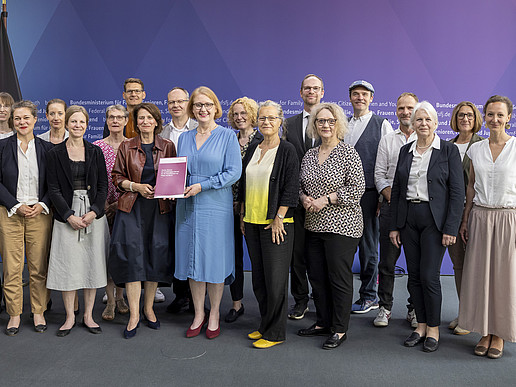 Beiratsmitglieder und die Familienministerin vor einer Pressewand im Bundestag stehen für ein Gruppenbild nebeneinander. Die Ministerin hält den Bericht in der Hand.