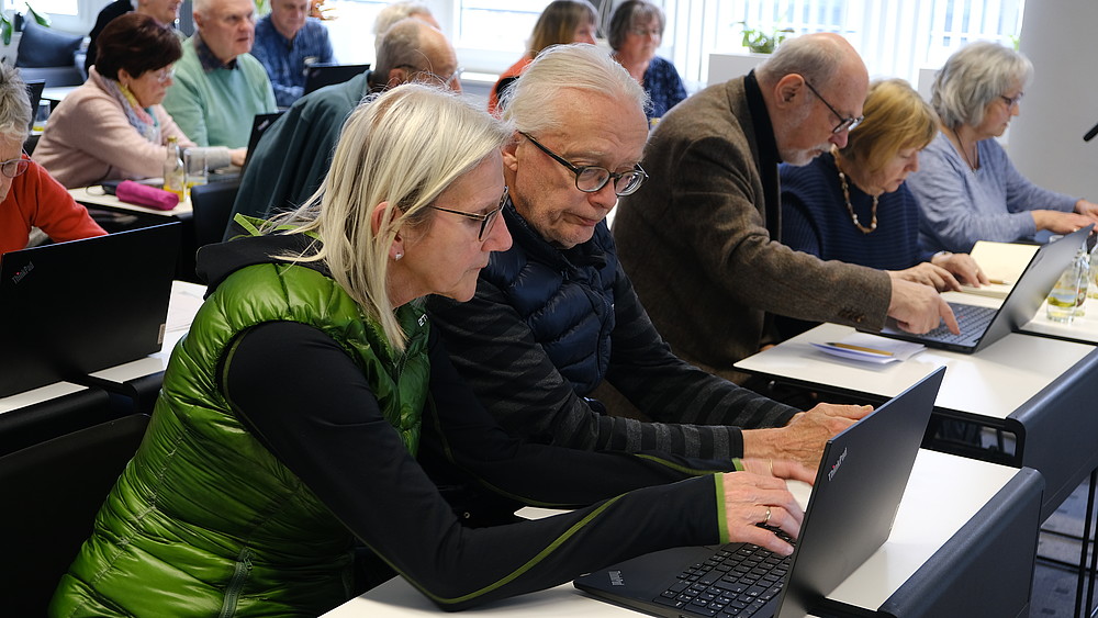 Eine Seniorin und ein Senior sitzen im Seminarraum umgeben von anderen Seniorinnen und Senioren und schauen auf einen Laptop.