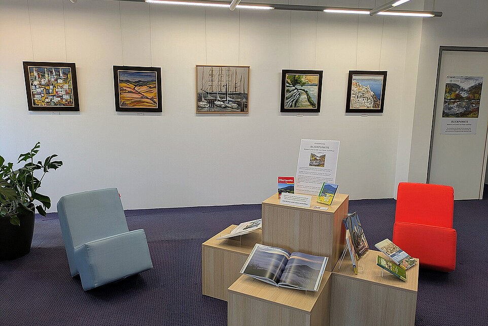 In einem Ausstellungsraum in der Hochschulbibliothek hängen Gemälde an der weißen Wand und im Vordergrund befinden sich moderne Sessel und Holzwürfel, auf denen Bücher präsentiert werden.