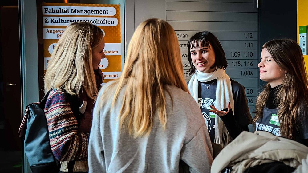 Vier Studieninteressierte stehen als Gruppe zusammen und unterhalten sich vor Informationstafeln der HSZG.
