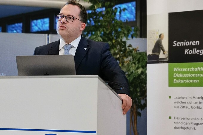 Prof. Kratzsch steht hinter einem Rednerpult und wendet sich an das Publikum. Im Hintergrund steht ein HSZG-Roll up.