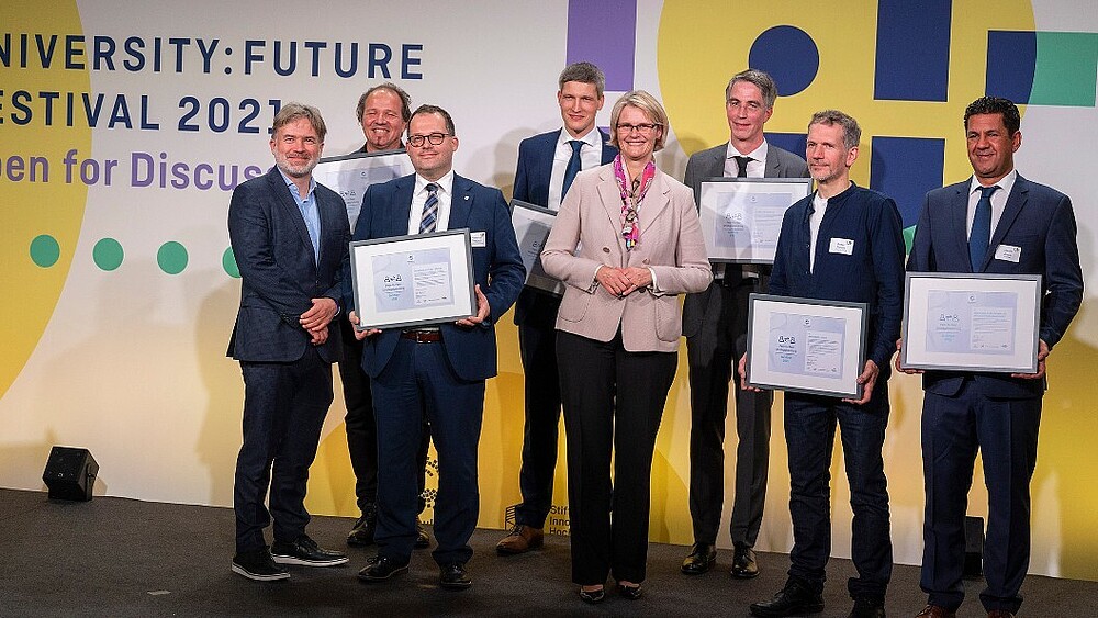Auf einem Podium stehen sieben Männer, mit einer Urkunde in der Hand, in der Mitte steht die Bundesbildungsministerin und lächelt in die Kamera.