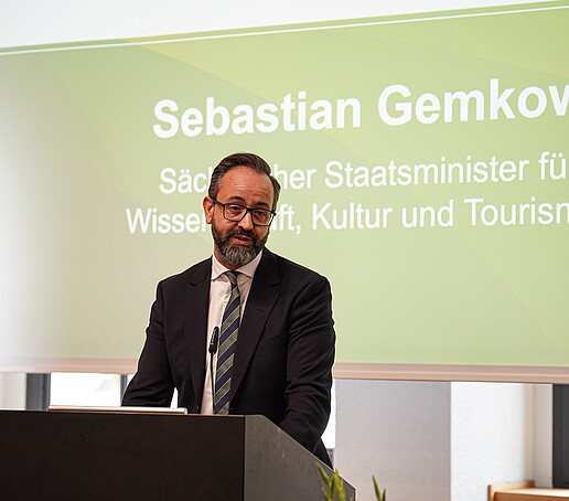 Ein Mann im Anzug mit Brille spricht an einem Rednerpult. Hinter ihm ist eine Präsentationsfolie mit seinem Namen und seiner Funktion als Wissenschaftsminister eingeblendet.