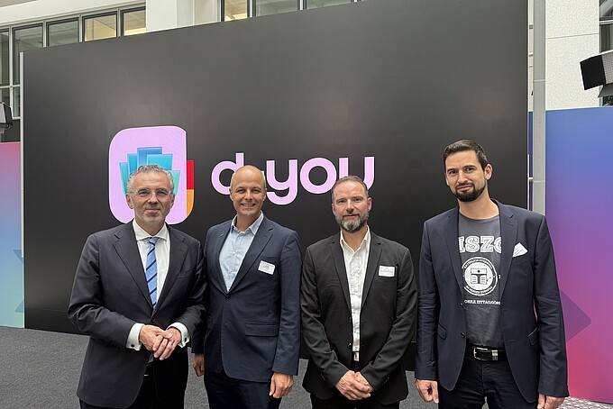 Vier Teilnehmer des Launchpartner-Treffens stehen vor einer großen Wand mit dem d-you-Logo.