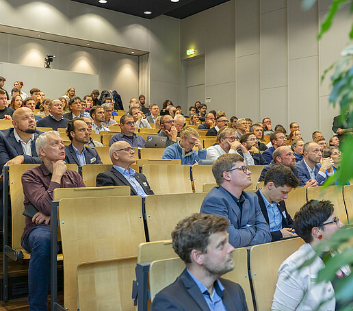 Die Sitzreihen sind mit Publikum besetzt, dass den Blick nach vorn richtet.