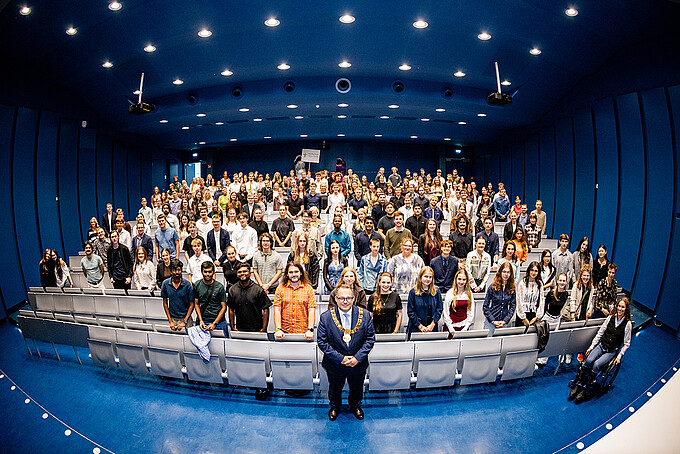 Große Gruppe von Erstsemestern im Audimax am Campus Zittau, vorne Rektor Prof. Dr.-Ing. Alexander Kratzsch; zwischen den Studierenden sind auch zwei als Dinosaurier verkleidete Personen zu sehen.