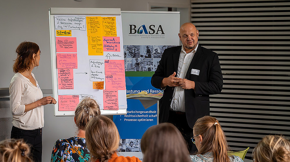 Ein Professor steht vor vollen Sitzreihen und erklärt etwas anhand eines Boards, das mit Zetteln bestückt ist.