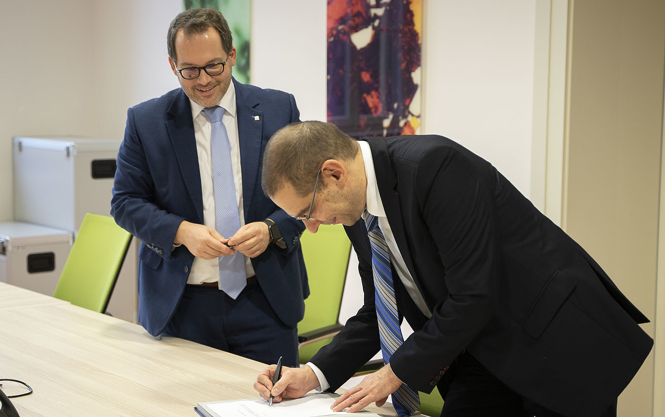 Dr. Zeller steht gebeugt über einem Tisch und unterzeichnet die Honorarprofessur-Urkunde. Der Rektor steht lächelt hinter ihm und beobachtet ihn dabei.