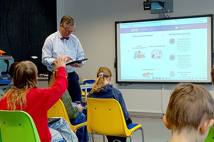 Prof. ten Hagen steht neben einem Bildschirm und schaut auf ein Tablet. Vor ihm sitzen Grundschüler auf Sitzreihen. Eine Schülerin hebt den Arm.