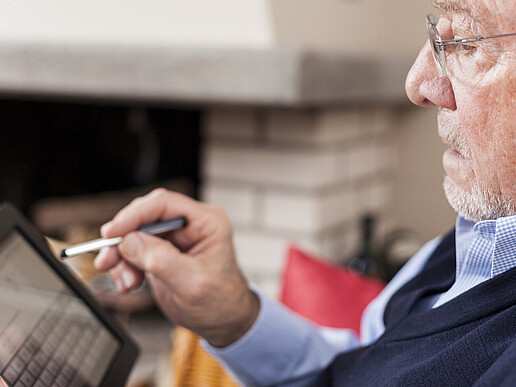 Ein älterer Herr mit Brille hält ein Tablet in einer Hand und einen Stift in der anderen Hand, mit dem er das Display bedient.