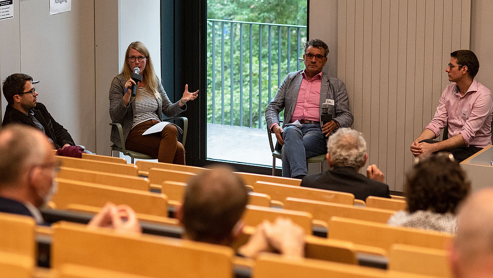 Vier Teilnehmende einer Podiumsdiskussion sitzen im Halbkreis, diskutieren und hören einander zu. Mehrere Teilnehmende sitzen in den Hörsaalrängen.