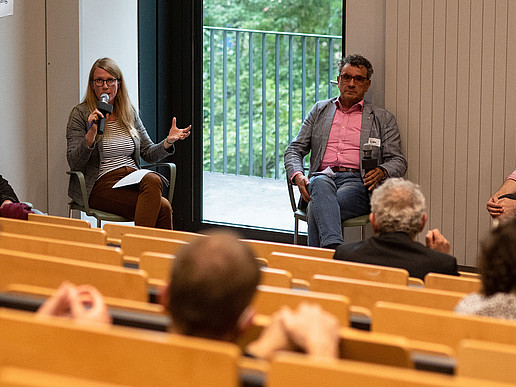 Vier Teilnehmende einer Podiumsdiskussion sitzen im Halbkreis, diskutieren und hören einander zu. Mehrere Teilnehmende sitzen in den Hörsaalrängen.